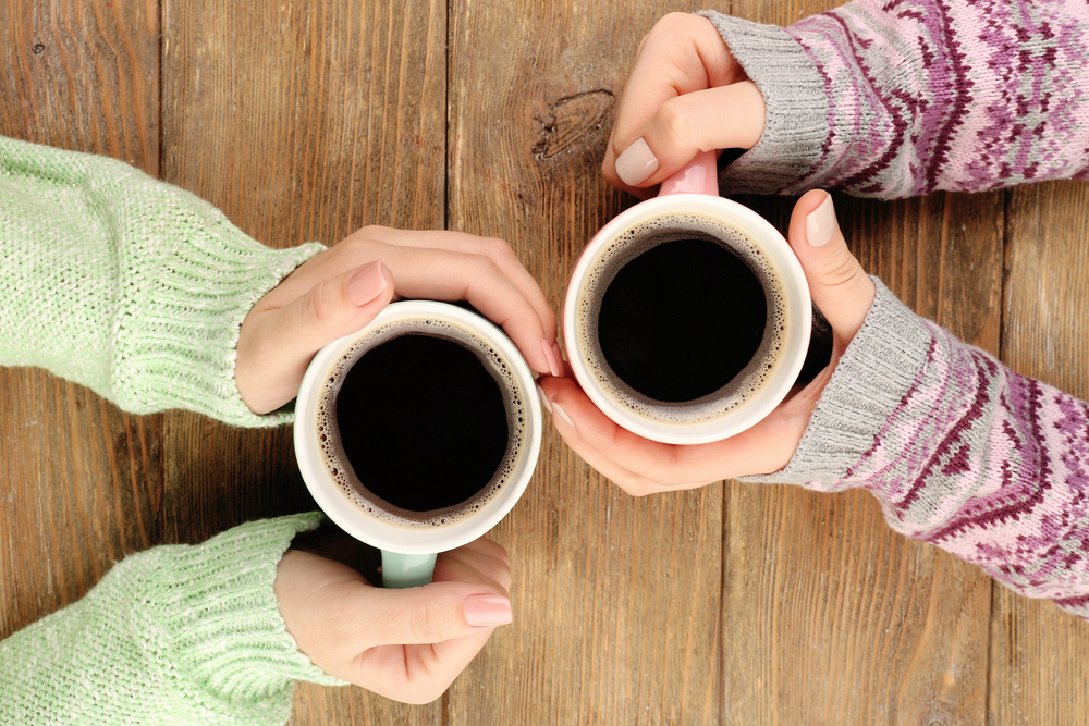 Female hands (wearing sweaters) holding cups of coffee on rustic wooden table
